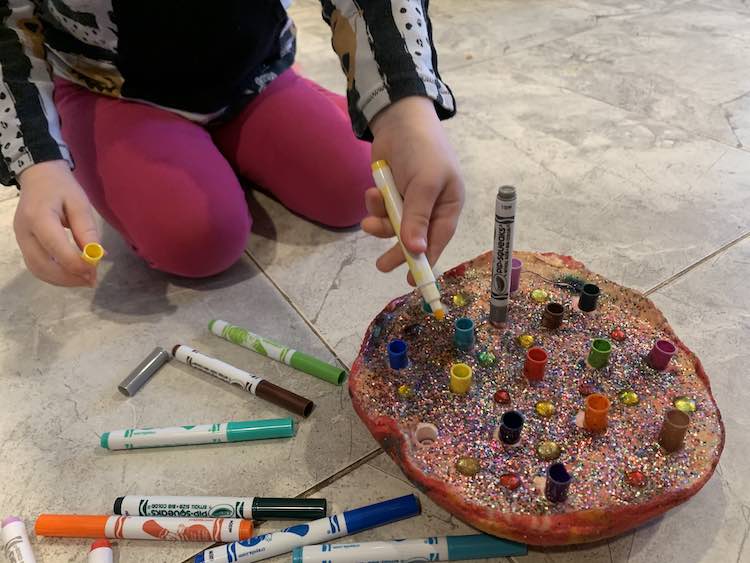 The felt pen marker has a single marker already in it while a yellow one is poised to be added. Zoey is on her knees behind the felt pen holder and there are various markers on the floor beside it.