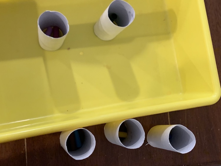 Image shows our yellow sensory bin with two vertical toilet paper rolls inside it and three below it. Inside of each roll are a couple marker lids.