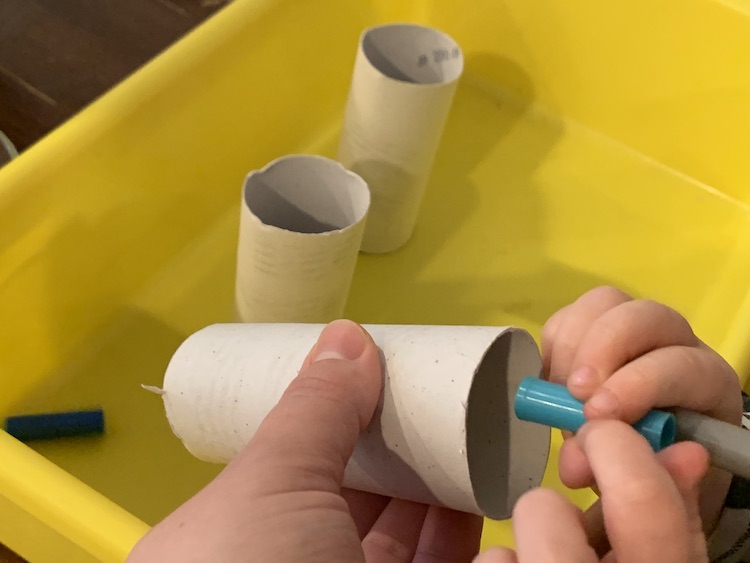 In the foreground I'm holding an angled toilet paper roll and Zoey is about to drop a turquoise lid into it. In the background there's a yellow bin with two vertical rolls and a lid that just came from the 'slide'.