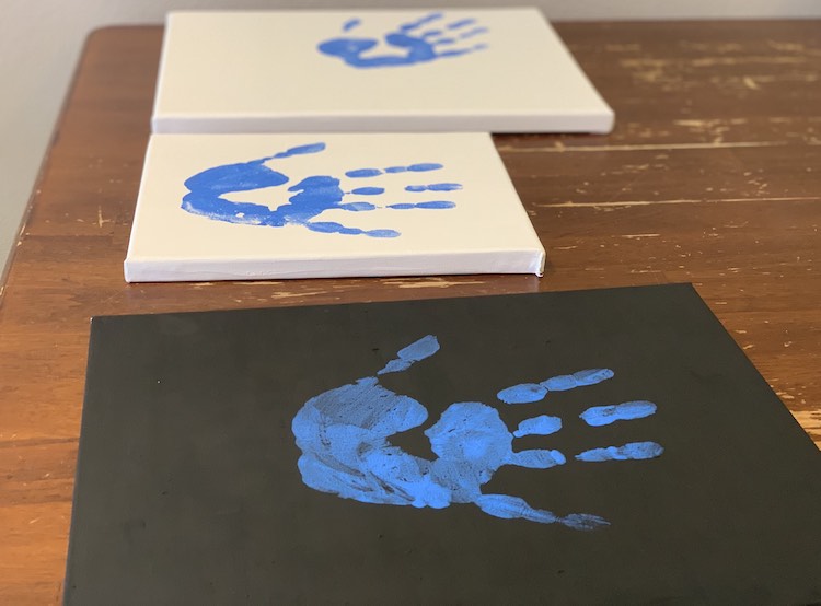 The three canvases laid out next to each other. In the image the canvases are turned sideways with the black one closest and the other two canvases beside it going further away. All three canvases have a blue handprint on it with the closest two centered in the middle and the furthest away one at an angle. 