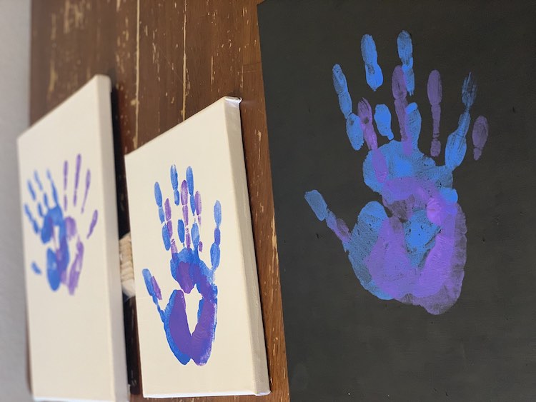 View of the the three paintings side by side on the table. The rightmost one has a black background and the blue and purple handprints are stacked over one another. The center small canvas is white with the prints, again, stacked. The far left and slightly blurry (in the image) is also white but just as large as the right canvas. Here the two handprints are slightly overlapping with the top of the hands leaning away from each other. 