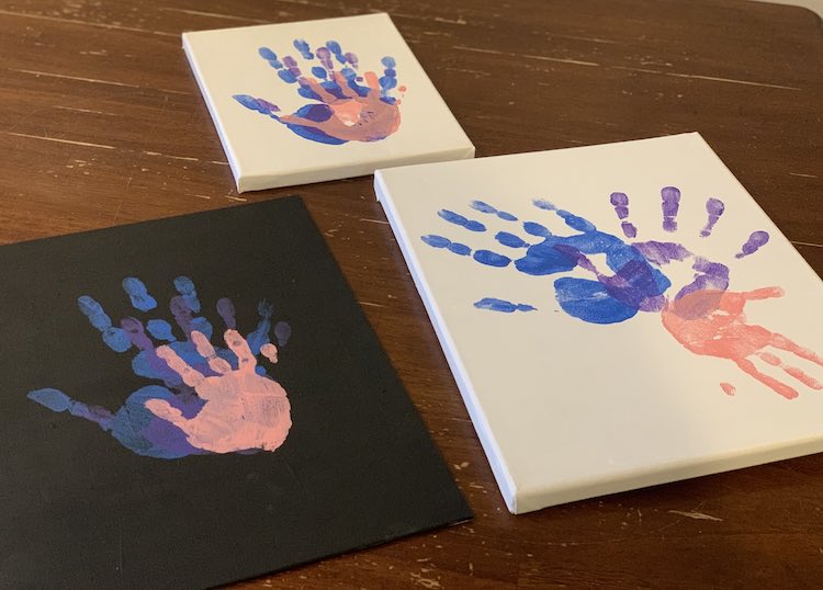 The three canvases are shown again with three handprints stamped on. The left (black) and right (white) canvases closer to the bottom of the photo are the larger ones while the smallest (white with stacked hands) is furthest away from the camera and is centered above the other two.