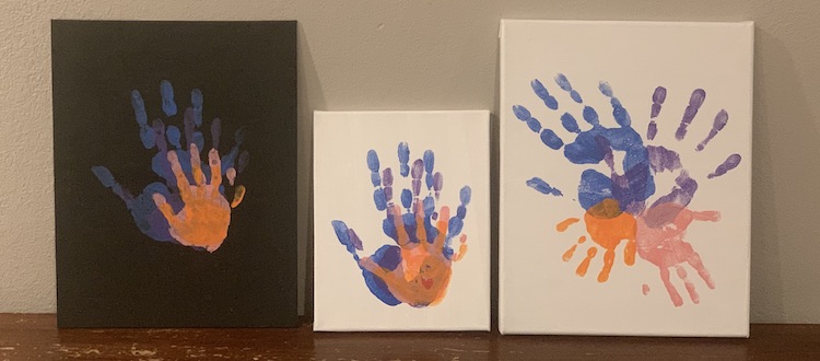 The three canvases leaning against the wall and sitting on the table. They're placed beside it each other so from left to right we have: 
1. the black canvas with the handprints stacked overtop of each other from biggest to smallest. 
2. The smallest canvas with the white background. The hands are the same color and are stacked like the left one. In the lower right corner of the palm there sits a red heart.
3. The white large canvas with the handprints forming an X with the largest handprints forming the top and the smallest ones forming the bottom legs of the X. 