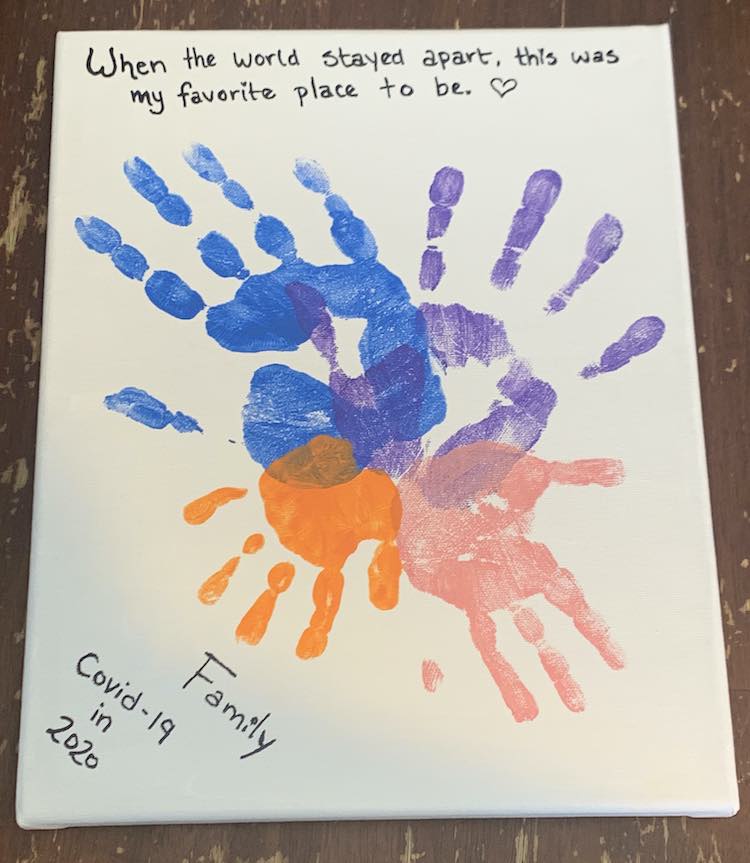 This canvas was painted white and the four handprints were stamped with the palms slightly overlapping with the fingers spread outward into a circle. The largest blue handprint is on the upper left side and then the handprints decrease in size going around until it reaches the smallest orange handprint in the lower left side. The other two handprints are purple (upper right) and pink (lower right). Above the handprints are the words "When the world stayed apart, this was my favorite place to be. <3". Below the hands several lines of text angled in the left corner reads "Family/Covid-19/in/2020".