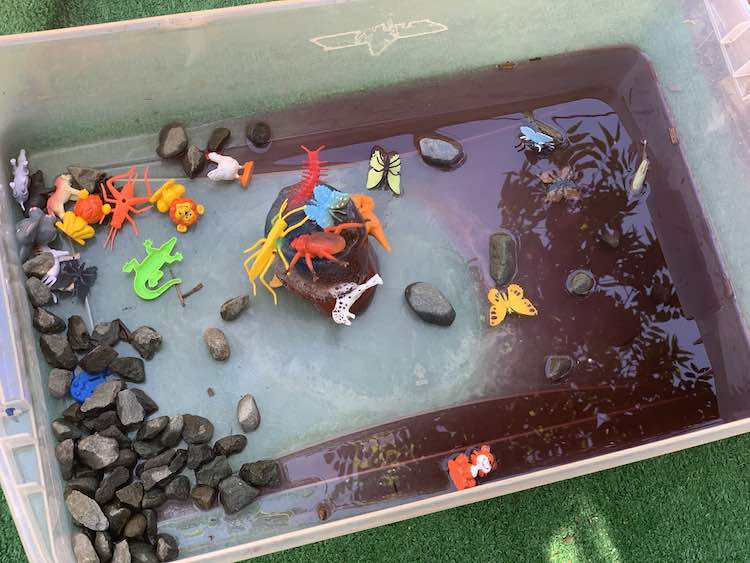 Overhead view of the bin showing the frozen chunk of ice in the center with plastic animals partially stuck within. The freed animals are scattered around with the purple-hued water to the right and rocks, mostly, to the left.