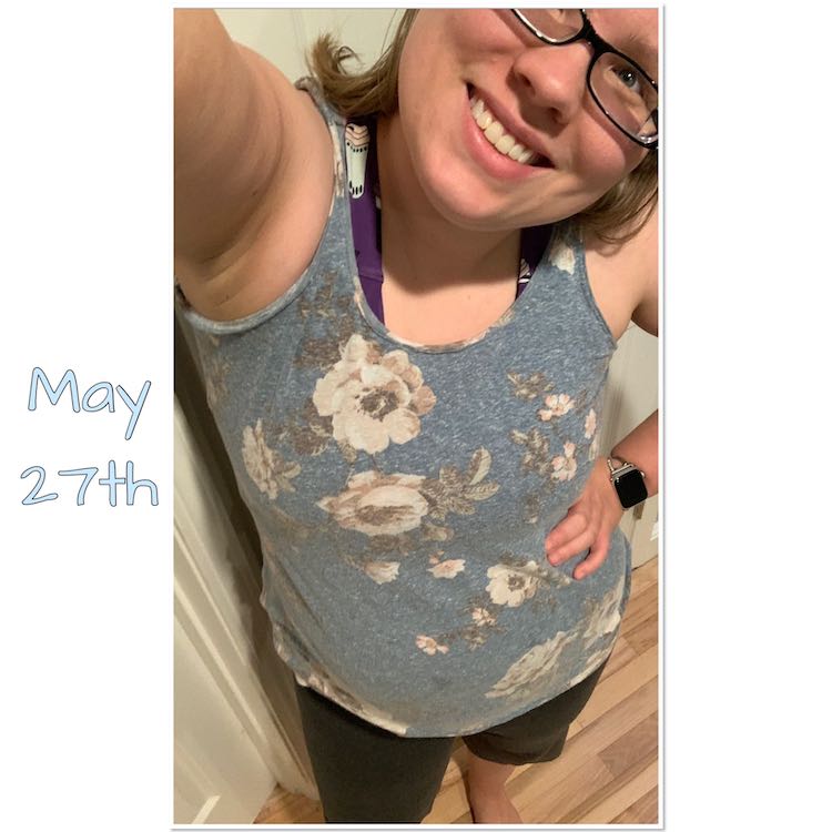 Selfie view of me tilting my head again so one of my eyes are visible. The arm not holding the phone is on my hips. I'm wearing a light blue tank with white flowers on it. Under the tank you can see the purple workout brazi and under the shirt you can see my grey pippa capris. 