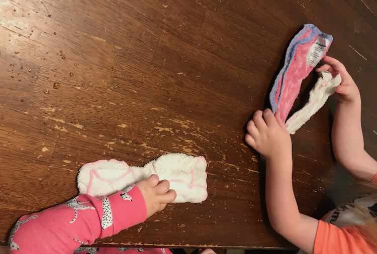 Overview of the table showing the kids arms holding washcloths. Zoey is visible with a single arm holding a washcloth. To the right Ada sits holding onto two washcloths. All the washcloths are folded over and frozen in place. 