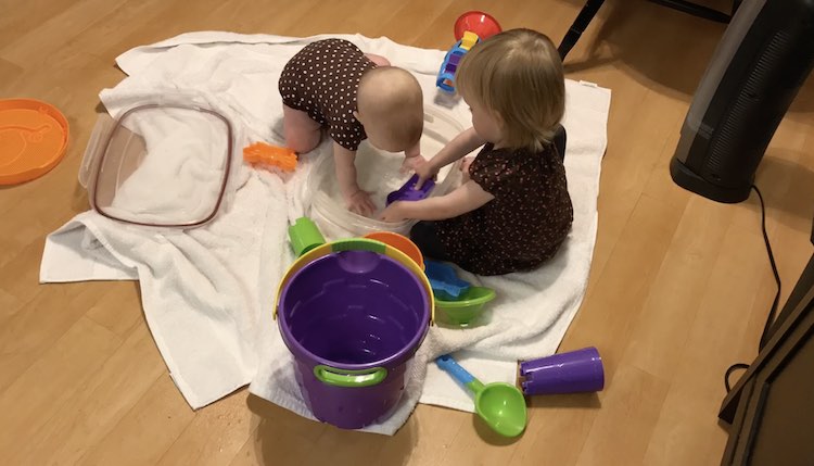 On the white towels on the ground are a clear bin with a baby playing while the toddler attempts to fill up her purple sand toy. Scattered around them are various other toys.
