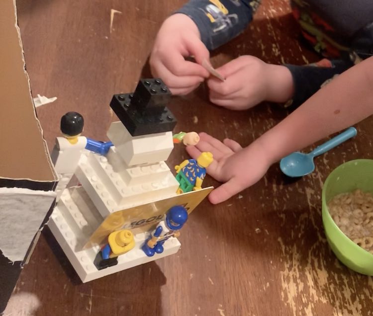 The back of the LEGO® snowman is showing with the one side partially hidden behind the corner of the box. You can see Zoey's hand behind the snowman as it props her up as she leans in closer (off photo). Behind her arm are Ada's hands holding the paper she pulled out of the advent calendar.