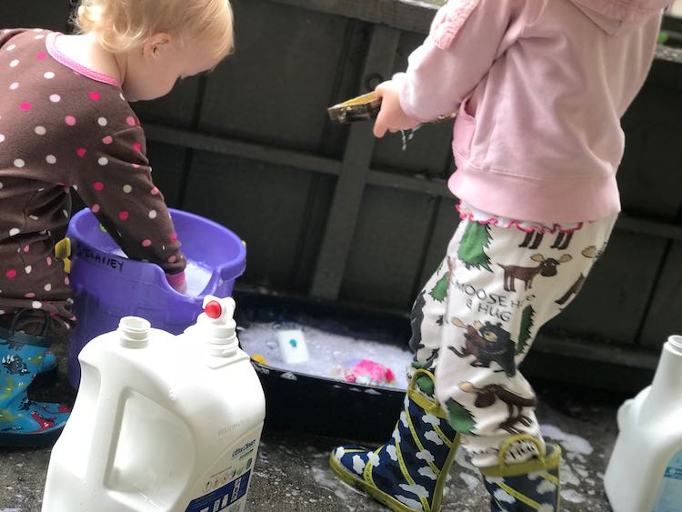 To the left Zoey leans over reaching into a purple sand pail filled with soapy water. To the right Ada walks towards Zoey carrying an item to the bin of soapy water. An emptied 