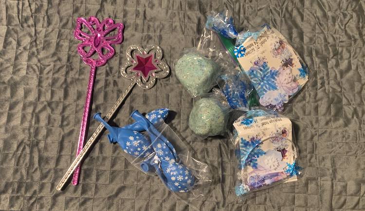 Top down view of two of the Melissa and Doug wands, a bag of blue snowflake latex balloons, two balls of gliittery blue playdough, and two clear cellophane bags with blue snowflake on it and Frozen items inside it. 