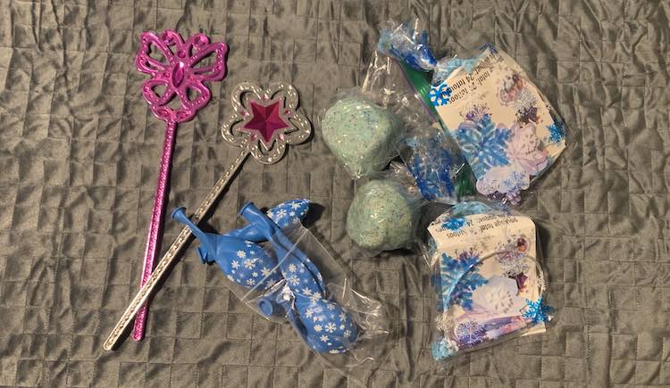Top down view of two of the Melissa and Doug wands, a bag of blue snowflake latex balloons, two balls of gliittery blue playdough, and two clear cellophane bags with blue snowflake on it and Frozen items inside it.