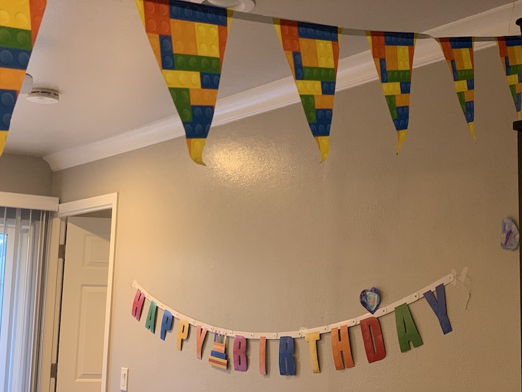 At the top of the image, close to the camera, the LEGO® banner hangs. In the background, on the wall, is the Happy Birthday banner with a LEGO® cake between the two words.