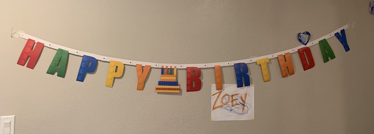 Closeup of the LEGO® Happy Birthday banner on the wall. There's masking tape on either end keeping it up. Over the d in Birthday one of Ada's older hearts hang. Under the IR in Birthday there's a doodled white sheet of paper with the word ZOEY in orange capital letters. 