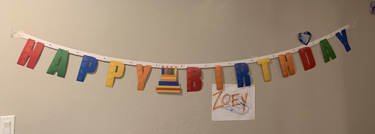 Closeup of the LEGO® Happy Birthday banner on the wall. There's masking tape on either end keeping it up. Over the d in Birthday one of Ada's older hearts hang. Under the IR in Birthday there's a doodled white sheet of paper with the word ZOEY in orange capital letters.