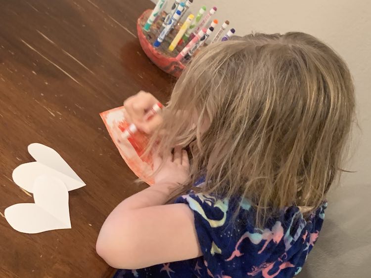 Side of Ada sitting at the table coloring a paper heart red. Behind her is holder with all the markers and in front of her are two more paper hearts.