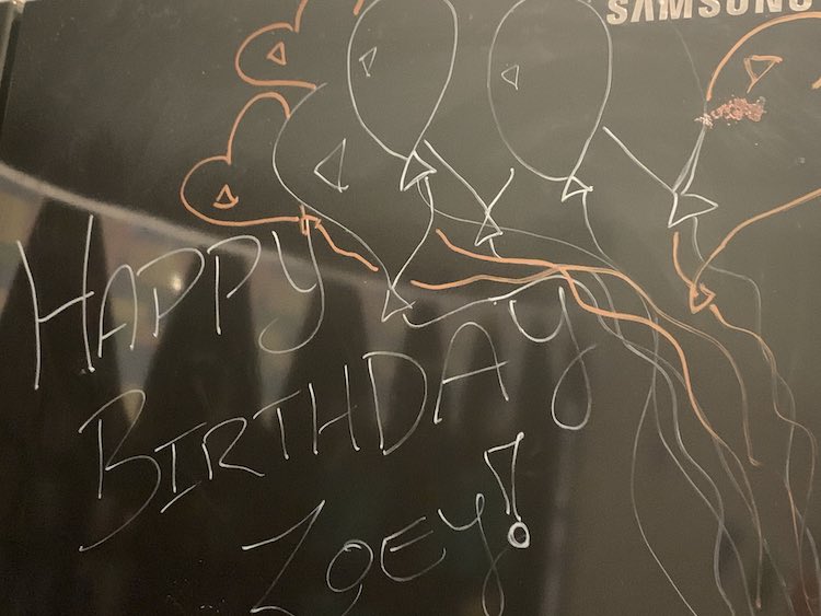 Black glossy fridge with "Happy Birthday Zoey!" in white along with white balloons and red heart balloons.