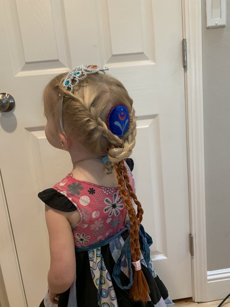 Photo showing Zoey's back with the dangling Anna braids. Her hair is french braided over the sides of the crown and then at the bottom the braids switch to normal and the Anna braids, with the blue centerpiece, is clipped between them. Zoey's braids are wrapped around the Anna braids and then pony-tailed in place so they're less visible.