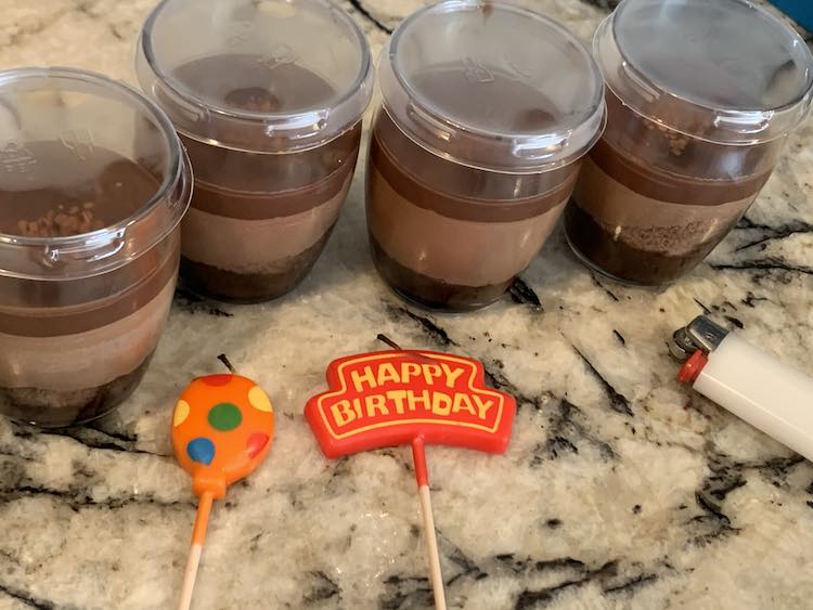 Closeup of four of the chocolate mousse from Costco. They beside each other with distinct layers in a glass cup, each, and a plastic lid. In front are two candles (one orange balloon and one Happy Birthday one) with a white lighter going off photo to the right.