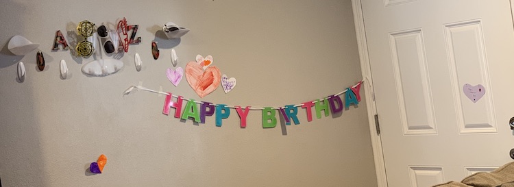 Wall with a multicolored sparkly Happy Birthday banner. There are six colored paper hearts around it and above it all, to the side, are the command hooks we use for the kids' jackets.