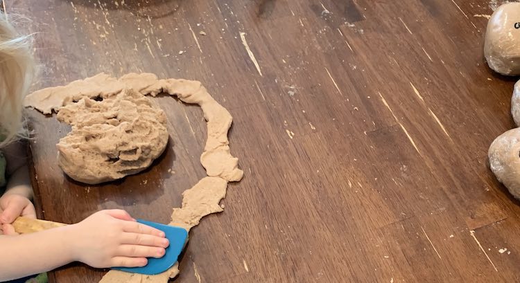 Image shows a ball of brown playdough with bits removed at the top. Around it is a squished path of playdough with Zoey leaned over squashing a section with a blue spatula. Off to the right you can see the edge of some plastic wrapped balls. 