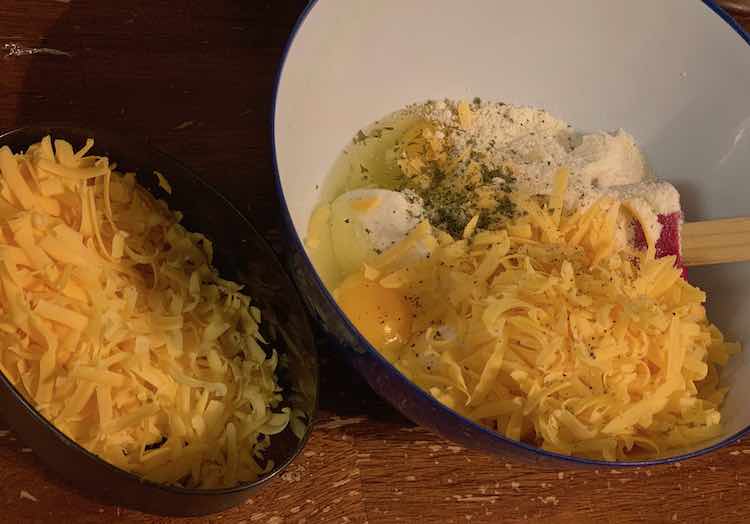 A large plastic bowl is on the right with cottage cheese, parmesan cheese, eggs, dried parsley, and grated cheddar cheese inside. There's a spatula resting inside the ingredients. To the left is an Ikea container with the extra grated cheese for on top.