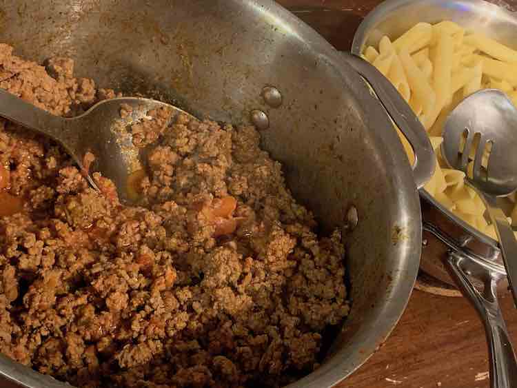 There are two pots in this image. On the left the large pot is half full with a ground beef and diced tomato mixture with a spoon resting inside. To the right is a smaller pot filled with penne pasta and a slotted spoon. 