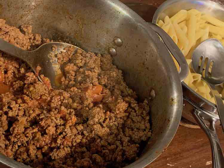 There are two pots in this image. On the left the large pot is half full with a ground beef and diced tomato mixture with a spoon resting inside. To the right is a smaller pot filled with penne pasta and a slotted spoon.