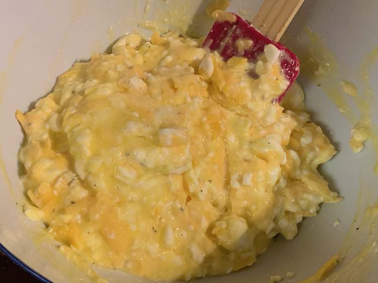 Closeup of the contents of the bowl showing the pink spatula inside a white, orange, and yellow mixture with the odd fleck of green (parsley). 