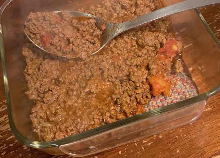 A square clear glass casserole dish sitting on a crocheted trivet. Meat sauce, including diced tomatoes, have been partially spread across the bottom. A metal spoon with meat sauce in it rests in the casserole dish. 