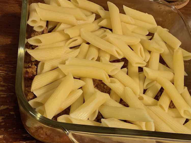 Closeup of the square glass casserole dish with penne noodles covering the meat sauce. You can see the bottom layer along the edge and between the noodles. 