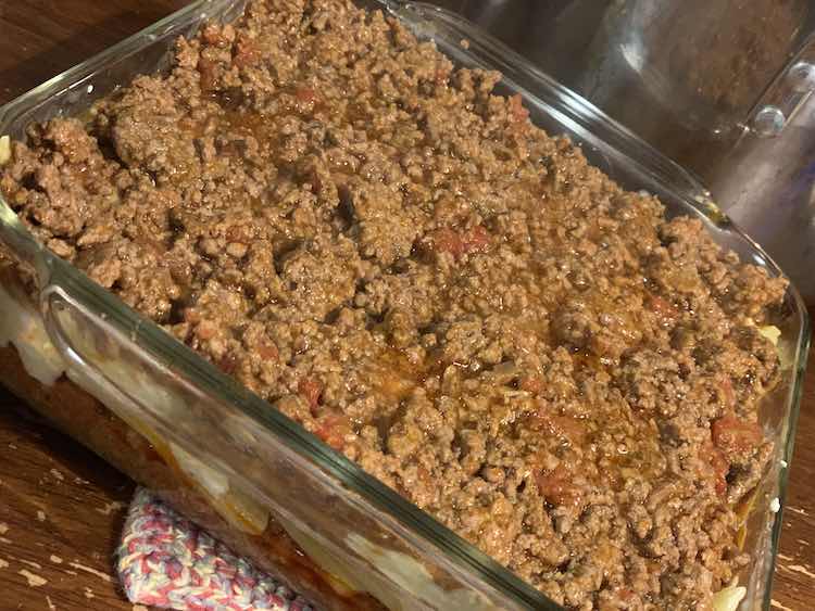 The clear casserole pan is mostly filled. The top is coated in meat and tomato mixture with a hint of pasta showing on the right side. From the front side you can vaguely see the pasta and cheese layer sandwiched between the two meat layers. 