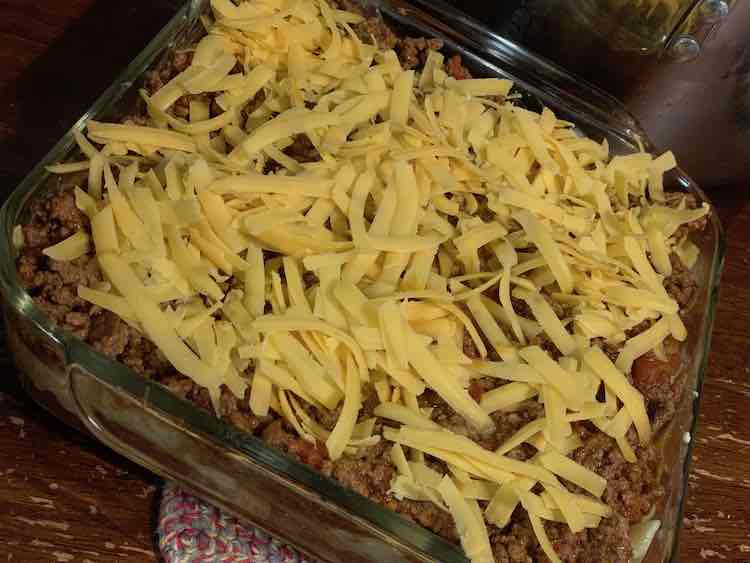 The glass clear casserole dish with a crocheted trivet underneath. The photo is mostly top down so it's harder to see the sides. The top shows the ground beef and tomato mixture covered in grated cheddar cheese with the odd pasta sticking out.