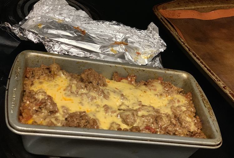 Overview of the top of a loaf pan filled with lasagna. The top is cheddar cheesy with ground beef showing through. Behind the loaf pan is the tinfoil that was on top, previously, and beside it is an empty lined cookie sheet.