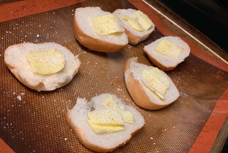 View, from above, of the orange Silpat lined cookie sheet with the bun halves on it and the slice of garlic sprinkled unmelted butter. 