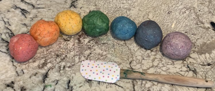 Image shows a row of sparkly colored balls of playdough and a spatula. The balls are red, orange, yellow, green, blue, black, and purple. 