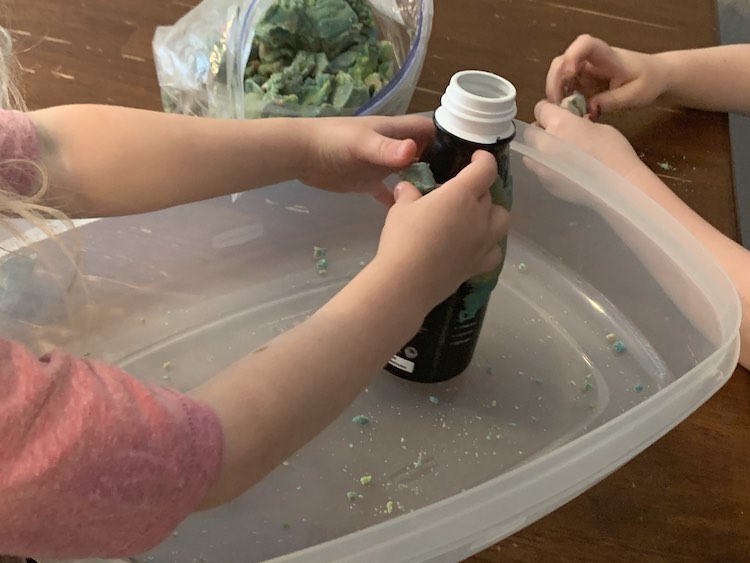 Image shows the bottle held by Zoey within a large container bottom as she presses a piece against the side. Ada is prepping a piece to put on after her and an opened bag of playdough sits behind. 