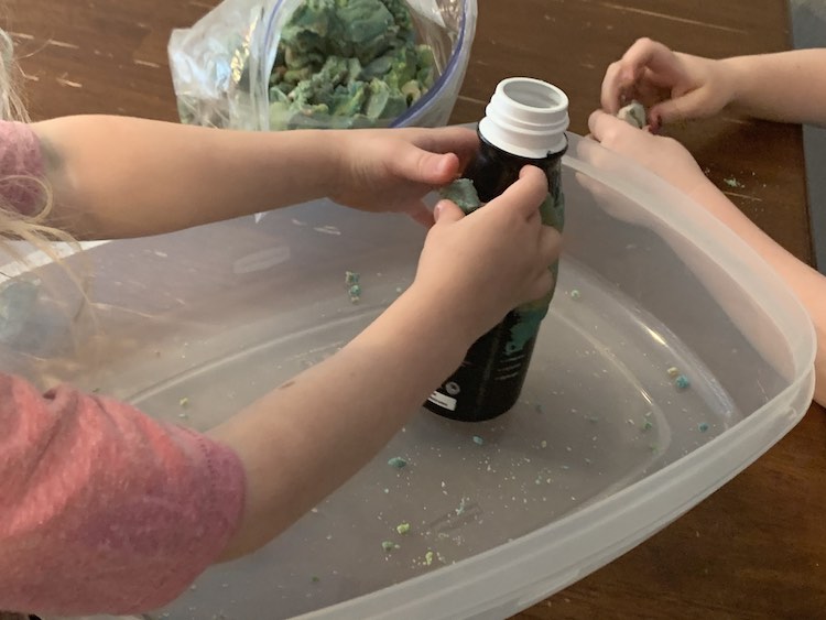 Image shows the bottle held by Zoey within a large container bottom as she presses a piece against the side. Ada is prepping a piece to put on after her and an opened bag of playdough sits behind.