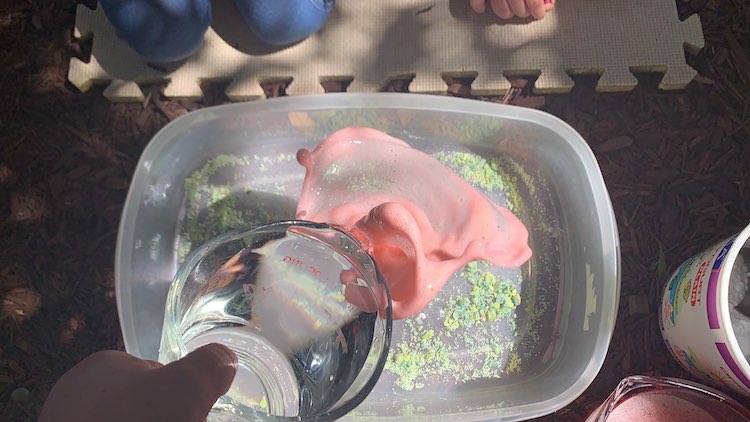 Image shows bubbly pink "lava" bursting out from the volcano. As the image is taken from above you can't see the volcano anymore although you can see the playdough coated floor of the bin and the part of the kids as they watch.