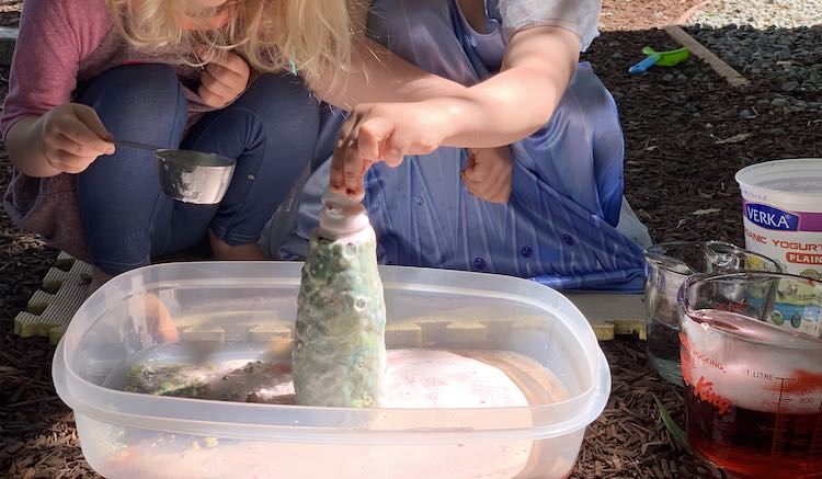 Image shows both kids crouched beside the bin with the bubbly wet volcano surrounded by lava remains. Zoey is holding the baking soda measuring cup and Ada is trying to put a ball of baking soda into the volcano.