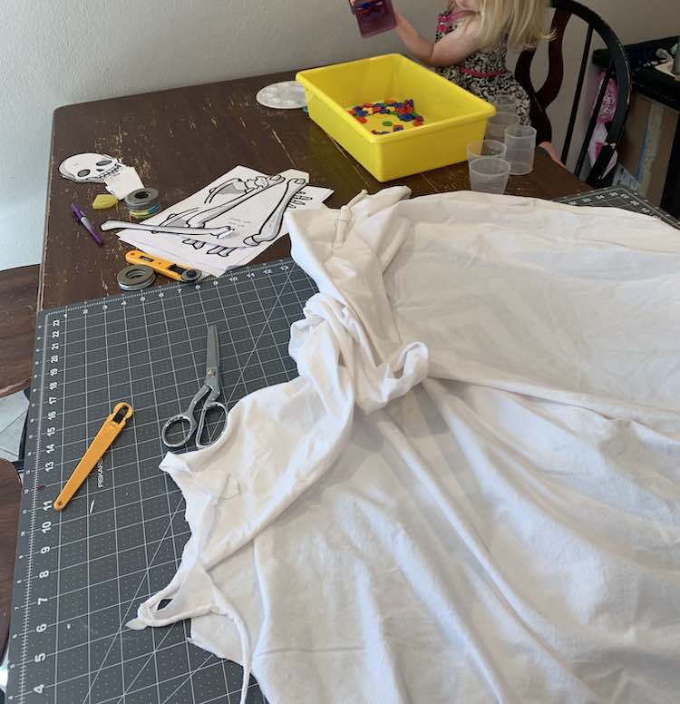 Image shows the table with my supplies: printed bones, white fabric, cutting mat, rotary cutter, and scissors laid out. In the background Zoey dumps out a vase-full of buttons.