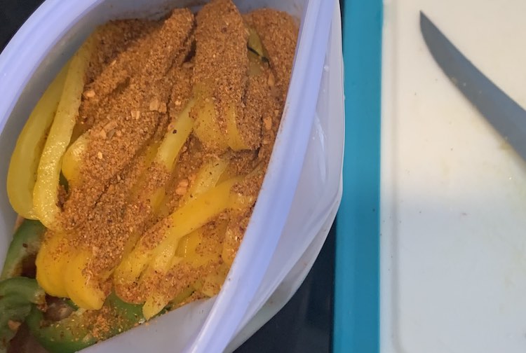 Image shows a view from above showing an opened blue Stasher bag. Inside you can see the seasonings sitting on top with yellow and then green pepper slices underneath. To the right is a plastic blue lined cutting board with part of a knife showing. 