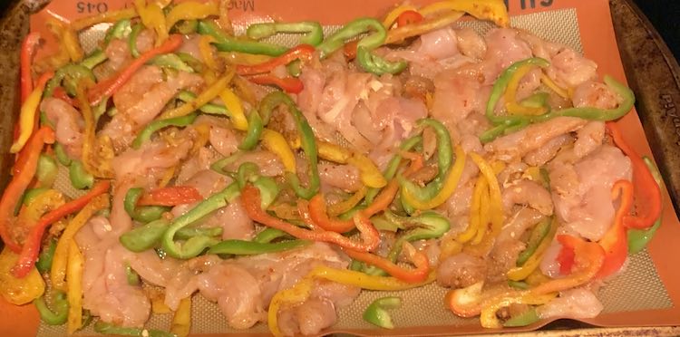 Closeup of the sheet pan showing raw pink chicken, sliced red peppers, sliced green peppers, and sliced onion.