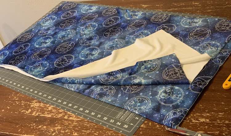 The fabric is folded in quarters, like the previous picture, but the top half is cut, lengthwise, up to the fold while a pair of sewing scissors sit poised to cut the other half.