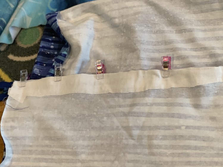 View of the wrong side of the striped fabric. The one end is folded up so it stretches horizontally across the image. There's a layer of interfacing along the edge and sewing clips along the top of it.