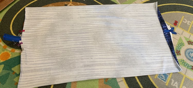The pillow is laid flat on a floor mat. The right side of the pillow is curling up showing the zipper underneath it. The left the of the pillow is clipped in the middle and the end of the zipper tape is sticking out from the side showing where the zipper is. 