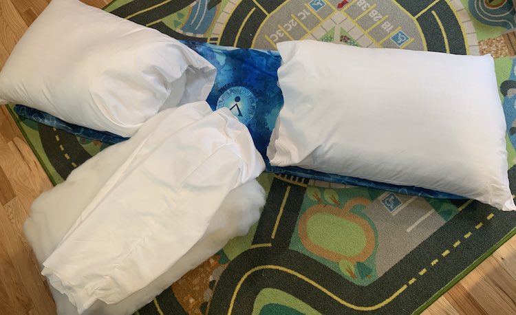 Image shows the long pillow cover laid out on the car mat mostly covered by the two pillows with the one end open. The center panel shows through but covering the bottom of it, angled up, lays the stuffing of the third pillow with it's pillow covering flopped overtop. 