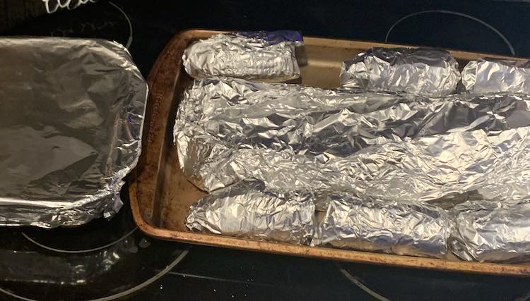 Closeup of the aluminun wrapped food sitting on the stovetop. To the left is a nine by nine casserole dish containing the beans, To the right is a sheet pan with the wrapped ribs down the center and the wrapped corn on the cob along either side. 