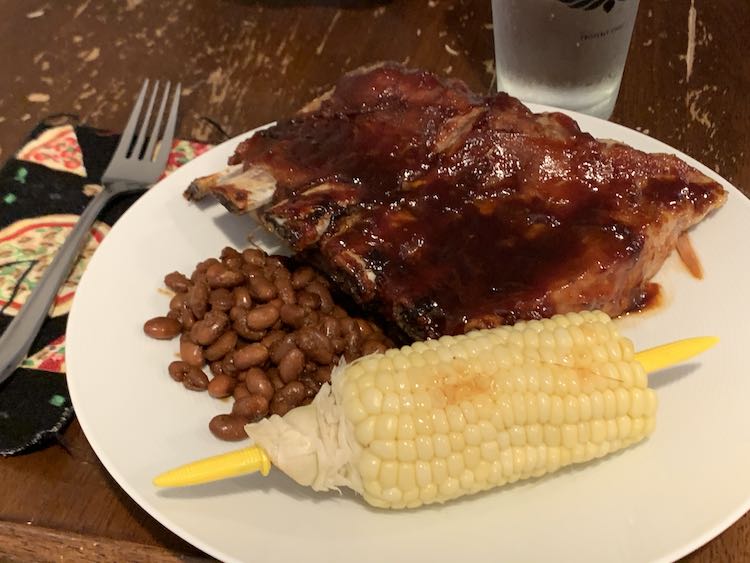 Image shows a white plat with sauced ribs, in the back, pinto beans, and slightly browned corn on the cob. To the left of the plate is a fork resting on a fabric napkin and a full glass of what sits behind everything. 