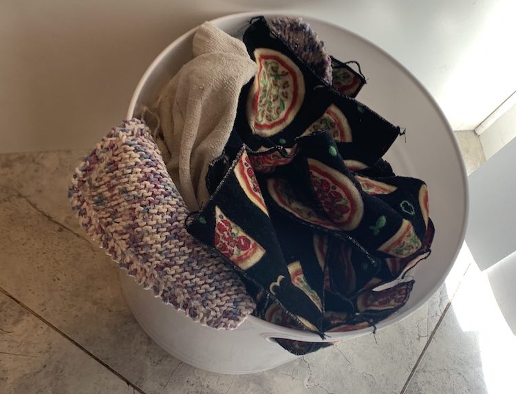 Overview of the white plastic pail in the corner of the kitchen between the cupboard side and blinds. Inside the pail yuo can see a couple dishcloths, a hand towel, and many pizza decorated napkins.